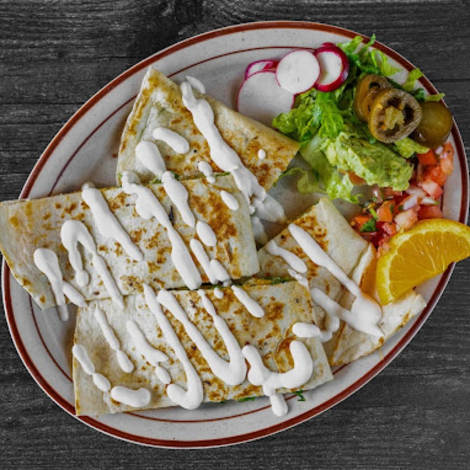 A plate of quesadillas drizzled with sour cream, served with guacamole, salsa, radishes, and jalapeños.